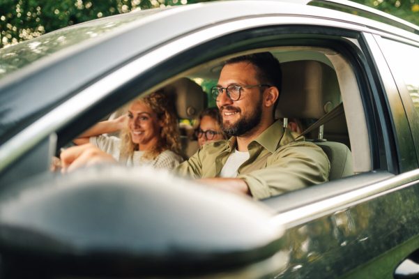 Portrait of a father driving a new car or having a test drive in the city while riding with his family. Parents and daughters spending vacation traveling by automobile and enjoying road trip.